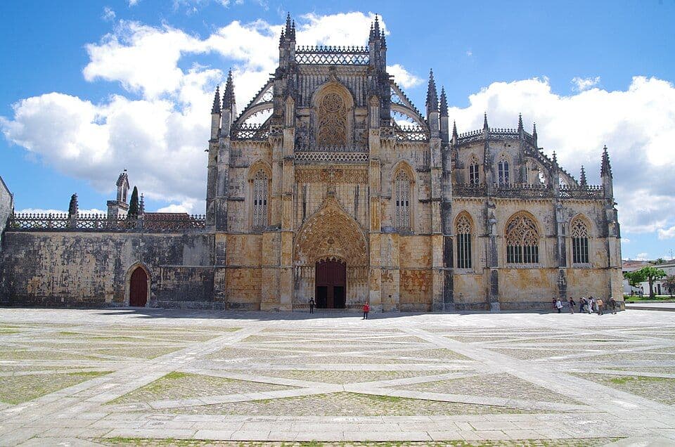 Fachada principal gótica do Mosteiro da Batalha, com o adro vazio e céu azul com nuvens