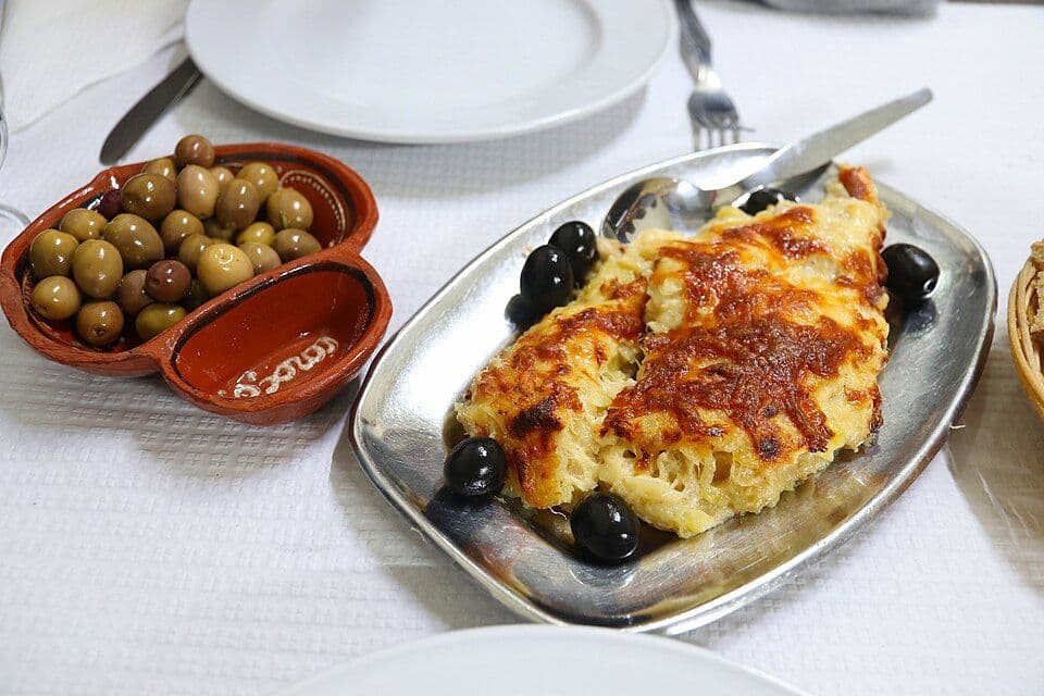 Travessa de bacalhau com natas gratinado com azeitonas pretas, ao lado de uma taça de azeitonas