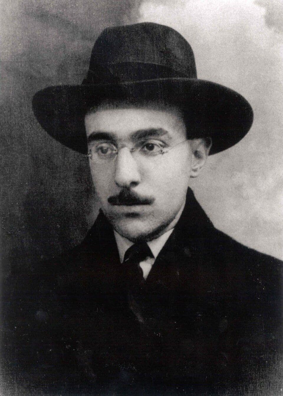 Fotografia a preto e branco de Fernando Pessoa jovem, de chapéu, óculos e bigode