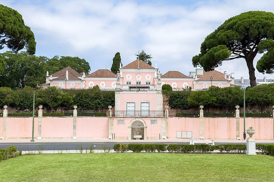 Palácio de Belém, cor-de-rosa, por detrás de um muro e de um relvado, entre pinheiros-mansos